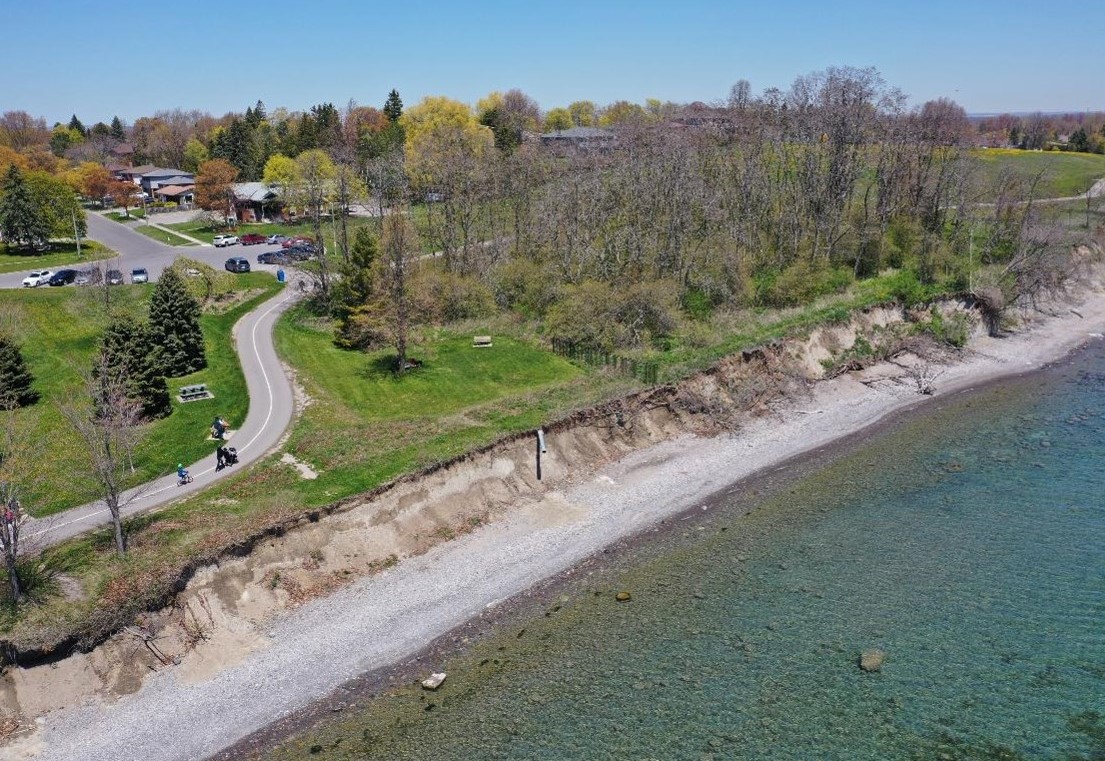 Aerial view of part of Reach 8, showing rocky/sandy shoreline and park area, with the Waterfront Trail running along the bluff and then around a treed area. There is an exposed pipe coming out of the eroded bluffs.