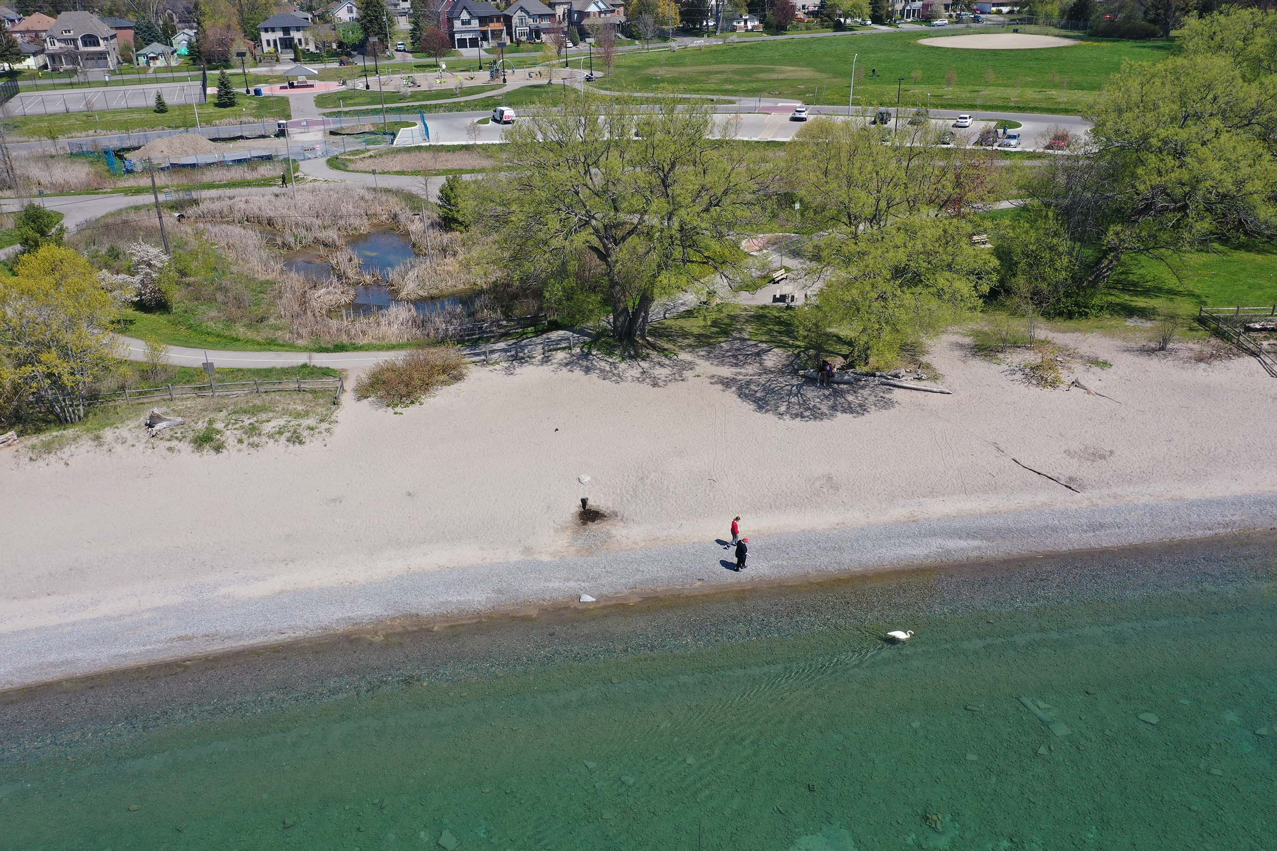 Aerial view of Paradise Beach taken from over Lake Ontario, with a parking lot and pond in the background. Two people are walking on the beach.
