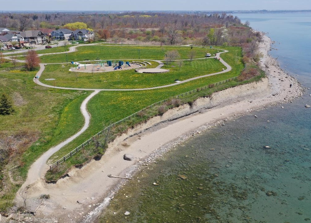 Aerial view of Reach 12 facing east, showing a large park space with a playground in the center, bordered by the Waterfront Trail. The shoreline is a narrow beach and has bluffs that gradually get higher further east. Audley Road S is in the background.