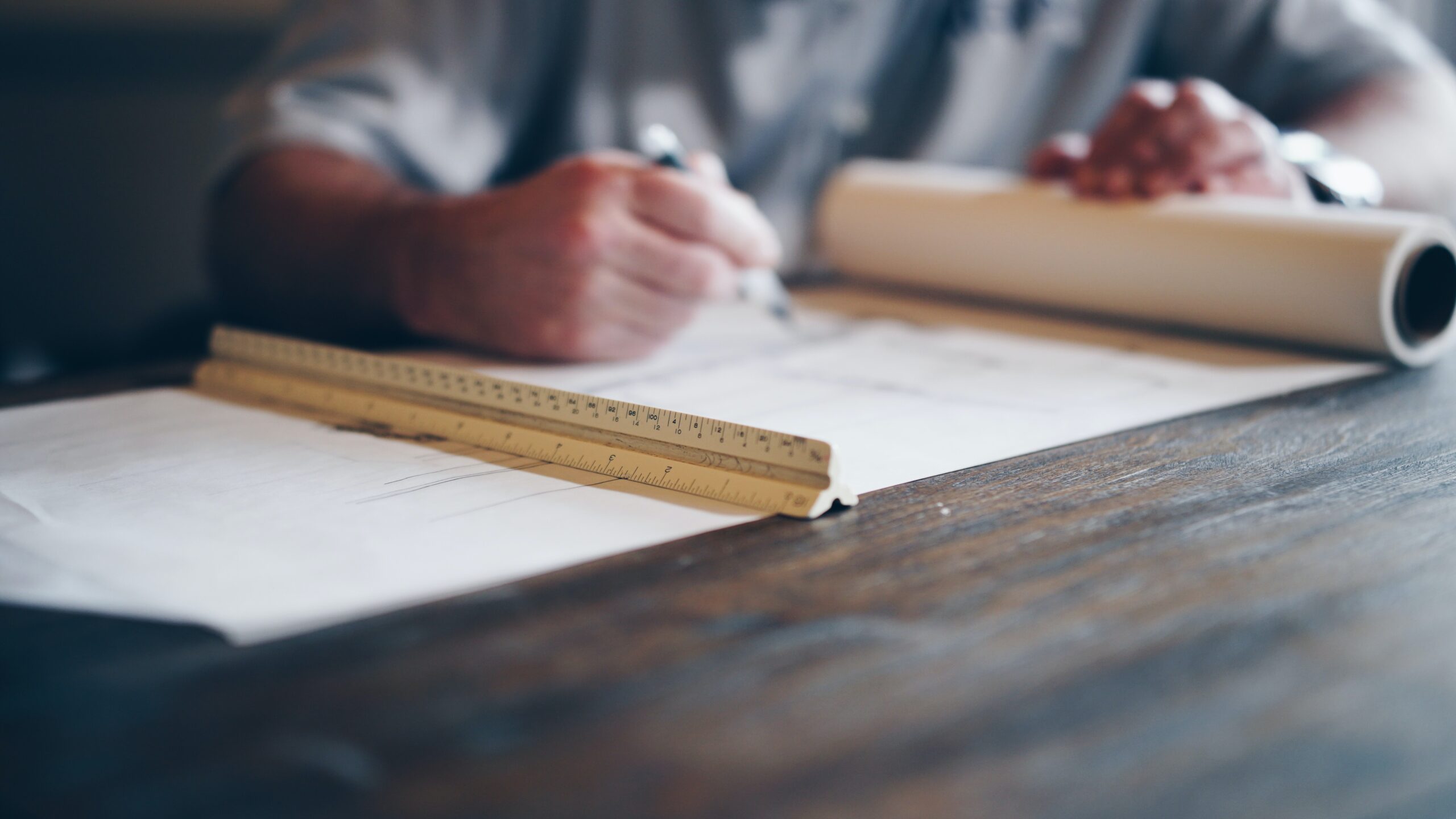 A photography of a man drafting a design drawing on a table, with a roll of paper and a ruler.