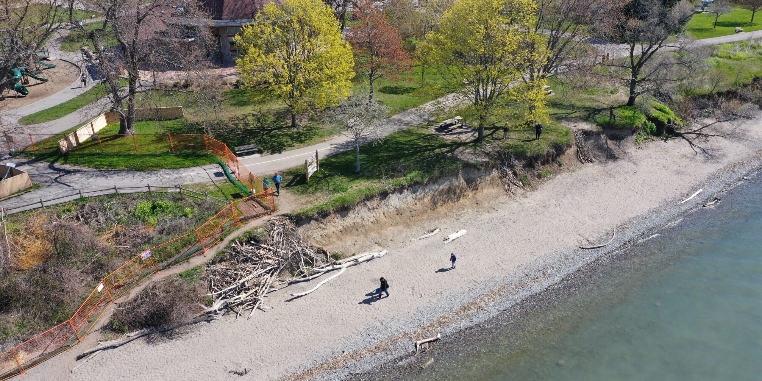 Aerial view of part of Reach 3, showing eroding bluffs close to the Waterfront Trail and other infrastructure. There is an orange fence on the bottom left corner closing one area, and several large tree limbs on the beach. Two people are walking on the beach.