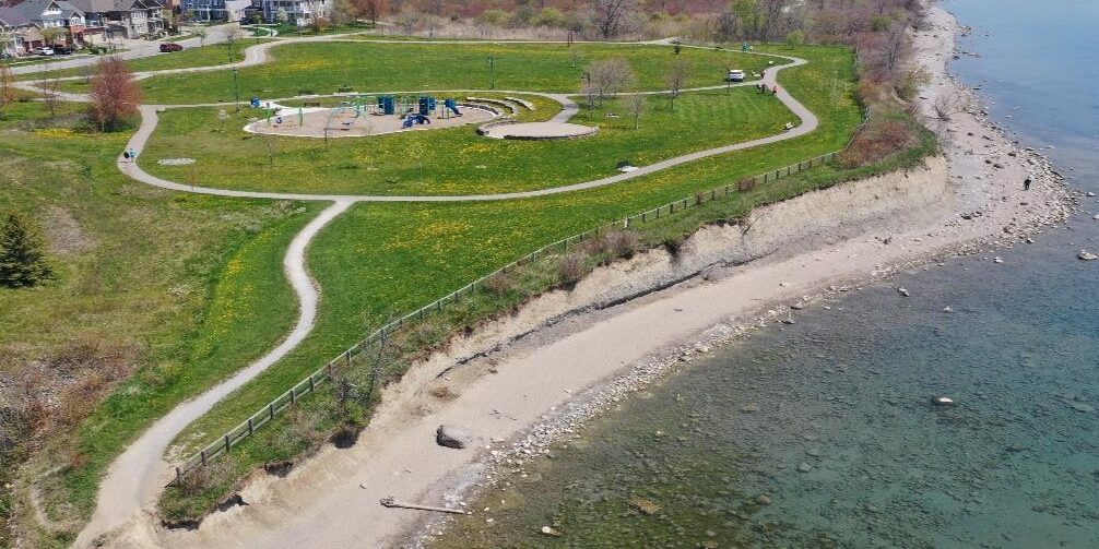 Aerial view of Reach 12 facing east, showing a large park space with a playground in the center, bordered by the Waterfront Trail. The shoreline is a narrow beach and has bluffs that gradually get higher further east. Audley Road S is in the background.
